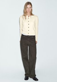 Cardigan beige clair avec six boutons noirs, associé à un pantalon large marron foncé. Le tissu semble doux avec une coupe décontractée.