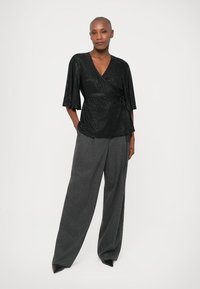 Femme portant un top enveloppant noir à manches trois-quarts et un large pantalon gris, se tenant avec les mains dans les poches sur fond blanc.