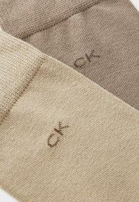 Šviesiai rudos ir kreminės spalvos medvilninės kojinės su briaunotu raštu ir logotipu „CK“ atitinkančiomis spalvomis viršuje. Minkšta tekstūra, vidutinio storio.