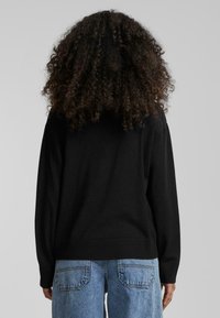 Pull noir avec ourlets et poignets côtelés, présentant une coupe décontractée. Assorti avec un pantalon en denim bleu clair, montrant l'arrière et la texture.