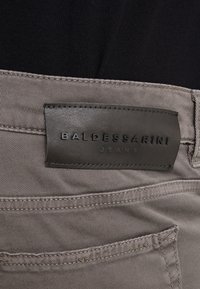 Acercamiento de la cinturilla de unos jeans grises con una etiqueta de cuero negro que dice "BALDESSARINI JEANS" cosida sobre el bolsillo trasero.