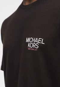 Μαύρο μπλουζάκι με λευκή επιγραφή "MICHAEL KORS" και μικρό ροζ κείμενο "NEW YORK CITY" στην αριστερή περιοχή του στήθους.