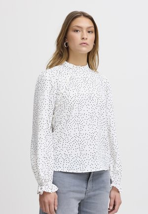 White long-sleeved blouse with black polka dots, featuring ruffled trim and buttons on one side, set against a neutral background.
