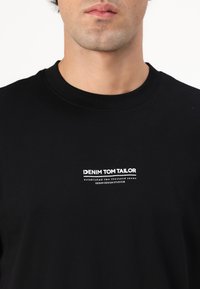 Sort bomulds t-shirt med rund hals og et trykt hvidt logo på brystet med teksten "DENIM TOM TAILOR." Simpelt design, afslappet pasform.
