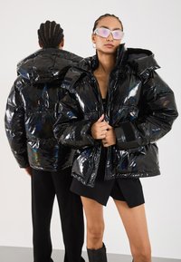 Black glossy puffer jacket with a hood, featuring a shiny finish and ribbed cuffs. Model wearing it, paired with black shorts and knee-high boots.