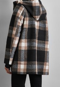 Rutig ullkappa i svart, brunt, vitt och grått. Har huva, långa ärmar och ribbade muddar. Mjuk struktur med avslappnad passform.