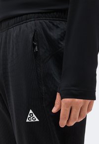 Gros plan sur la main d'une personne à côté d'un pantalon de sport noir texturé avec un petit logo blanc ACG près de la fermeture éclair de la poche à la ceinture.