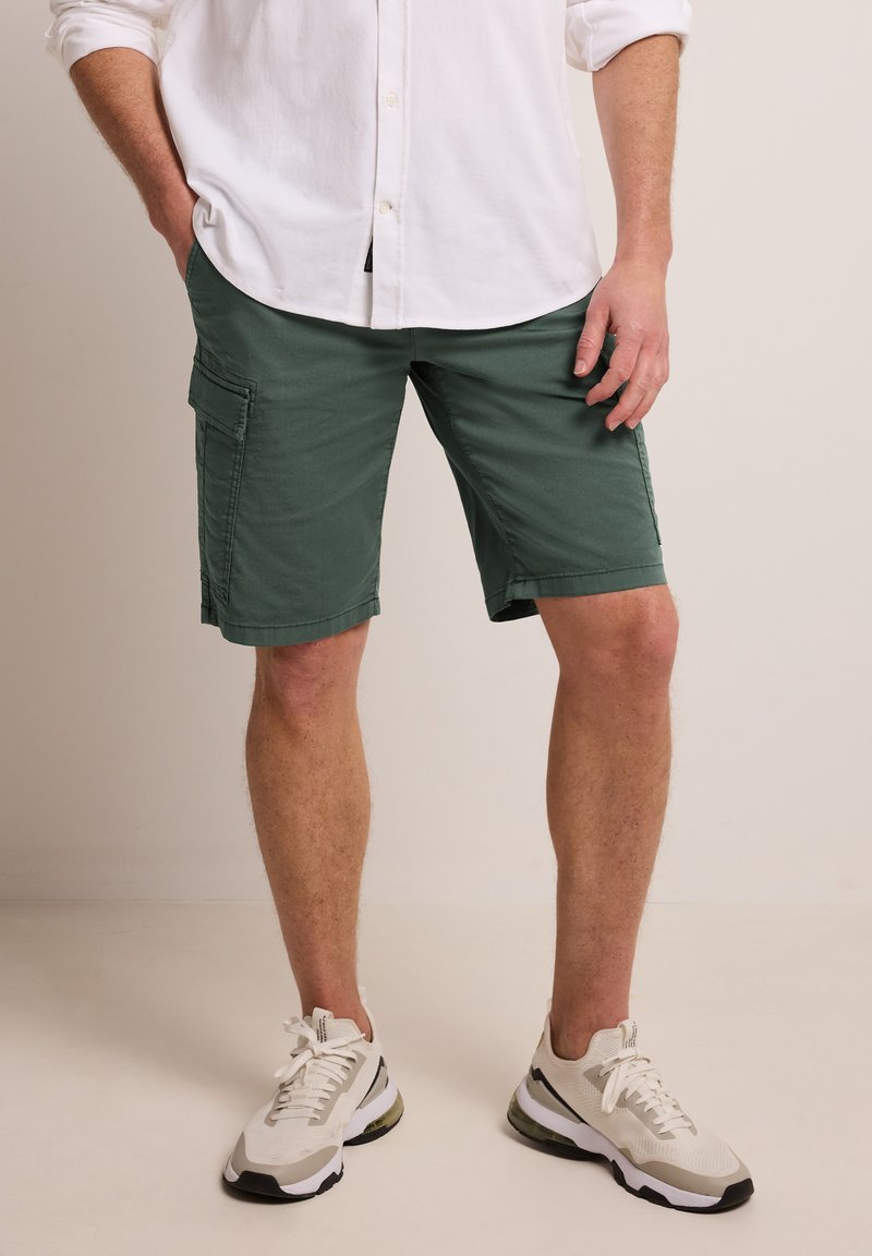 Homme portant un short cargo vert, une chemise blanche à boutons et des baskets blanches, debout avec une main dans la poche devant un fond uni.