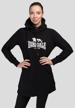 Kvinde stående med hænderne på hoften iført en sort Lonsdale London hættetrøje med en løvelogo på en ensartet lys grå baggrund.
