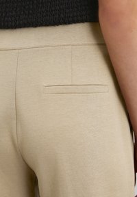 Pantaloni beige con una texture liscia, caratterizzati da una tasca a filetto sul retro e una vita aderente. Design minimalista con linee pulite.