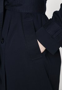 Tmavě modrý kabát s páskem, zapínáním na knoflíky a bočními kapsami. Látka má hladkou texturu s jemnými šicími detaily.