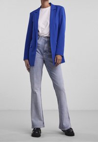 Persona vistiendo un blazer azul brillante, camiseta blanca, jeans de cintura alta y acampanados de lavado claro, y zapatos de plataforma gruesa negros frente a un fondo sencillo.