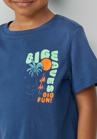 Marineblaue Baumwoll-T-Shirt mit einem bunten Grafikdesign und dem Text "GROßE WELLEN, GROßER SPASS!" sowie Illustrationen von einer Palme, einer Sonne und einem Surfbrett.