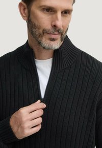 Maglione a coste nere con colletto alto e mezza zip, indossato sopra una t-shirt bianca; presenta un tessuto testurizzato e un design aderente.