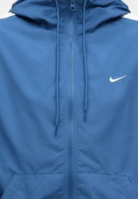 Kék kapucnis pulóver elülső cipzárral, húzózsinórokkal, kenguruzsebekkel és kicsi, fehér Nike swoosh logóval a mellrészen.