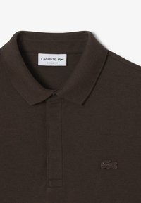 Brun polo skjorta i texturerad tyg, med klassisk krage, knappslå och en broderad logotyp på vänster bröst.