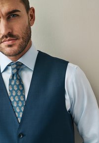 Marineblauer Weste über einem hellblauen Hemd. Die Krawatte hat ein Paisleymuster in beigen und blauen Tönen. Glatte Textur, taillierte Passform.