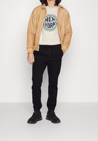 Giacca color bronzo sopra una t-shirt crema con grafica 1 NEW YORK 7, abbinata a pantaloni jogger neri e scarpe sportive nere. Trame di tessuto lisce.