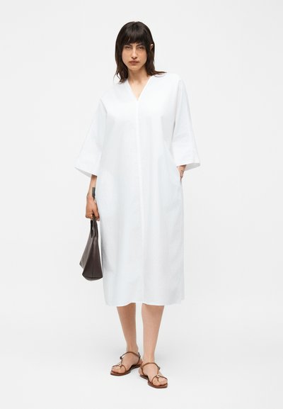 Γυναίκα που φοράει άνετο λευκό μίντι φόρεμα με μανίκια τριών τετάρτων, καφέ σανδάλια και κρατά μια μικρή σκούρα καφέ τσάντα.