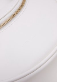 Bílá kožená taška se zipem a vyraženým logem. Hladký povrch s zaobleným tvarem a minimalistickým designem. Zlatý akcent na zipu.