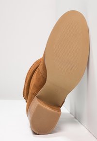 Bruna mockasinanklar har en slät gummisula med riller. Klacken är blockformad, vilket ger stabilitet och en avslappnad design.