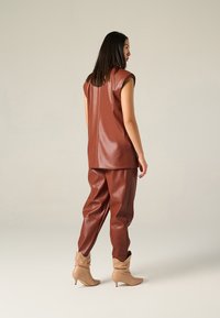 Top senza maniche in finta pelle marrone e pantaloni abbinati, caratterizzati da una vestibilità rilassata e una texture liscia. Stivaletti beige alla caviglia completano l'outfit.