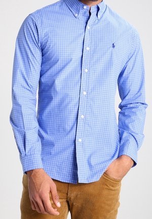 Hombre con una camisa de cuadros azul claro de manga larga abotonada con un pequeño logo bordado en azul oscuro y pantalones de pana marrones.