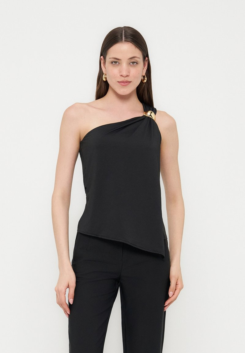 Γυναίκα που φοράει μαύρο τοπ με έναν ώμο και χρυσή λεπτομέρεια δαχτυλιδιού, καθώς και μαύρο παντελόνι, στέκεται μπροστά σε απλό φωτεινό φόντο.