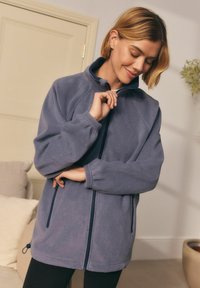Fleece jack in zacht grijs met een hoge kraag, volledige ritssluiting en zijzakken. Heeft elastische manchetten en een trekkoord onderaan voor aanpassing.