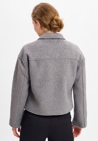 Szara kurtka z wełnianego materiału o teksturowanym wykończeniu, kołnierzem i opadłymi ramionami. Krótkie fason i zaokrąglony dół. Prosty, funkcjonalny styl.