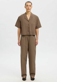 Braunes, kurzes, kurzärmliges Hemd mit Tasche, kombiniert mit locker sitzenden braunen Hosen. Die Textur wirkt glatt. Einfaches Design, neutrale Farbtöne.