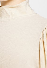 Vaaleanbeige, teksturoitu korkea kauluspaita, jossa on hillityt poimut olkapäillä ja pehmeä, kevyt kangas, joka laskeutuu vapaasti.