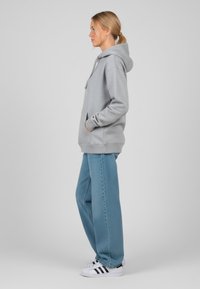 Felpa grigia con tasca frontale e cappuccio con coulisse, abbinata a jeans a gamba larga blu chiaro e sneakers bianche con strisce nere.