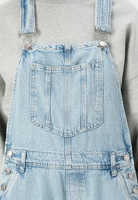 Salopette in denim azzurro chiaro con bottoni in argento e bretelle regolabili; presenta una tasca sul petto e un design classico da salopette.