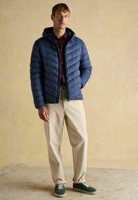 Marineblå pufferjakke med horisontale quiltninger, gråternet skjorte, cremede corduroybukser og grønne sneakers. Modellen står afslappet med hænderne i lommerne.