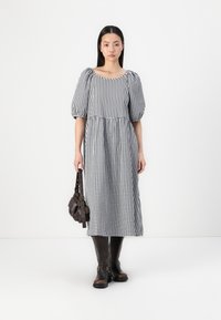 Vestido midi em padrão gingham preto e branco, com mangas bufantes, decote quadrado e cintura ajustada. A modelo segura uma bolsa castanha escura e usa botas castanhas.
