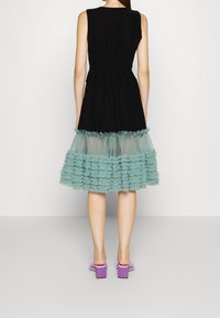 Robe noire sans manches avec un corsage froncé, un ourlet volanté en tulle transparent bleu sarcelle et des sandales à talons blocs violettes. Tissu lisse et texture superposée.