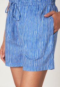 Pantaloni corti verticali a righe blu e bianche con vita elastica e coulisse annodata, mano appoggiata nella tasca laterale.