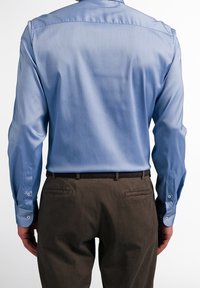 Uomo con camicia azzurra a maniche lunghe infilata in pantaloni marrone scuro, visto da dietro su sfondo bianco.