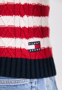 Rød og hvit stripete strikkegenser med et strukturert mønster, med en ribbet marineblå kant og et svart merke med "TOMMY JEANS."