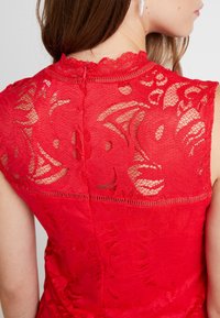 Primer plano de la espalda de una mujer que lleva un vestido de encaje rojo sin mangas con una cremallera y un delicado escote con forma de ondas.