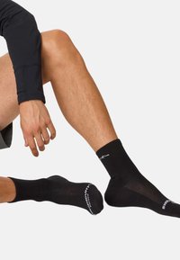Schwarze Sportsocken mit geripptem Bündchen und Mesh-Details. Versehen mit weißem Branding an der Seite und Logo an der Ferse, weiche Textur.
