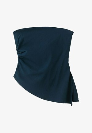 Žena s kučeravými vlasmi, oblečená v čiernom asymetrickom tubovom topu a svetlomodrých džínsoch s širokými nohavicami a vlnitými švovými detailmi, stojaca pred bielym pozadím.