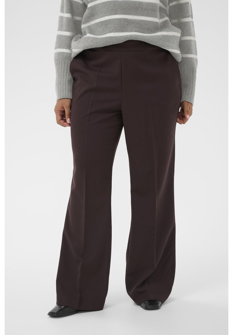 Femme portant un pantalon large marron foncé, un pull rayé gris et blanc et des chaussures pointues noires, debout devant un fond uni.
