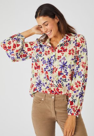 Bloes met bloemenpatroon op een lichte crèmekleurige achtergrond met rode, paarse en blauwe bloemen. Het heeft een kraag en pofferige mouwen.