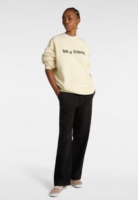 Sudadera crema con texto "dile a un amigo" en negro, combinada con pantalones anchos negros y zapatillas beige. Tejido suave, corte relajado.