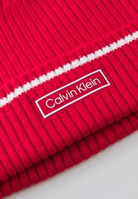 Rode gebreide beanie met een ribbelstructuur, voorzien van een witte streep en een rechthoekig Calvin Klein-label in witte letters op een rode achtergrond.
