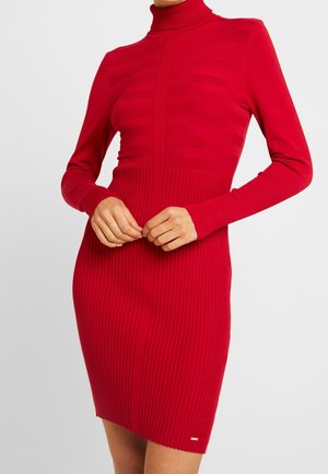 Femme portant une robe rouge côtelée ajustée, à manches longues et col roulé, les mains délicatement jointes à la taille.