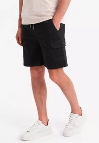 Shorts cargo noirs avec poches latérales, en tissu mélangé de coton. Portés avec un T-shirt crème clair à motifs et des baskets blanches.