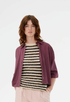 Kvinde med skulderlangt brunt hår iført en sort og beige stribet strikbluse, en åben blommefarvet skjorte og lyserøde bukser.